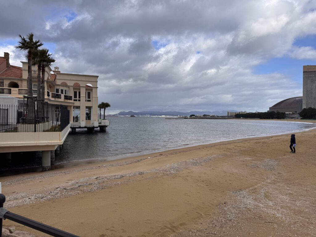  A peaceful beach scene with the same building as in image 1, but now viewed from a slightly different angle, showing a person walking along the beach with a cloudy sky in the background.