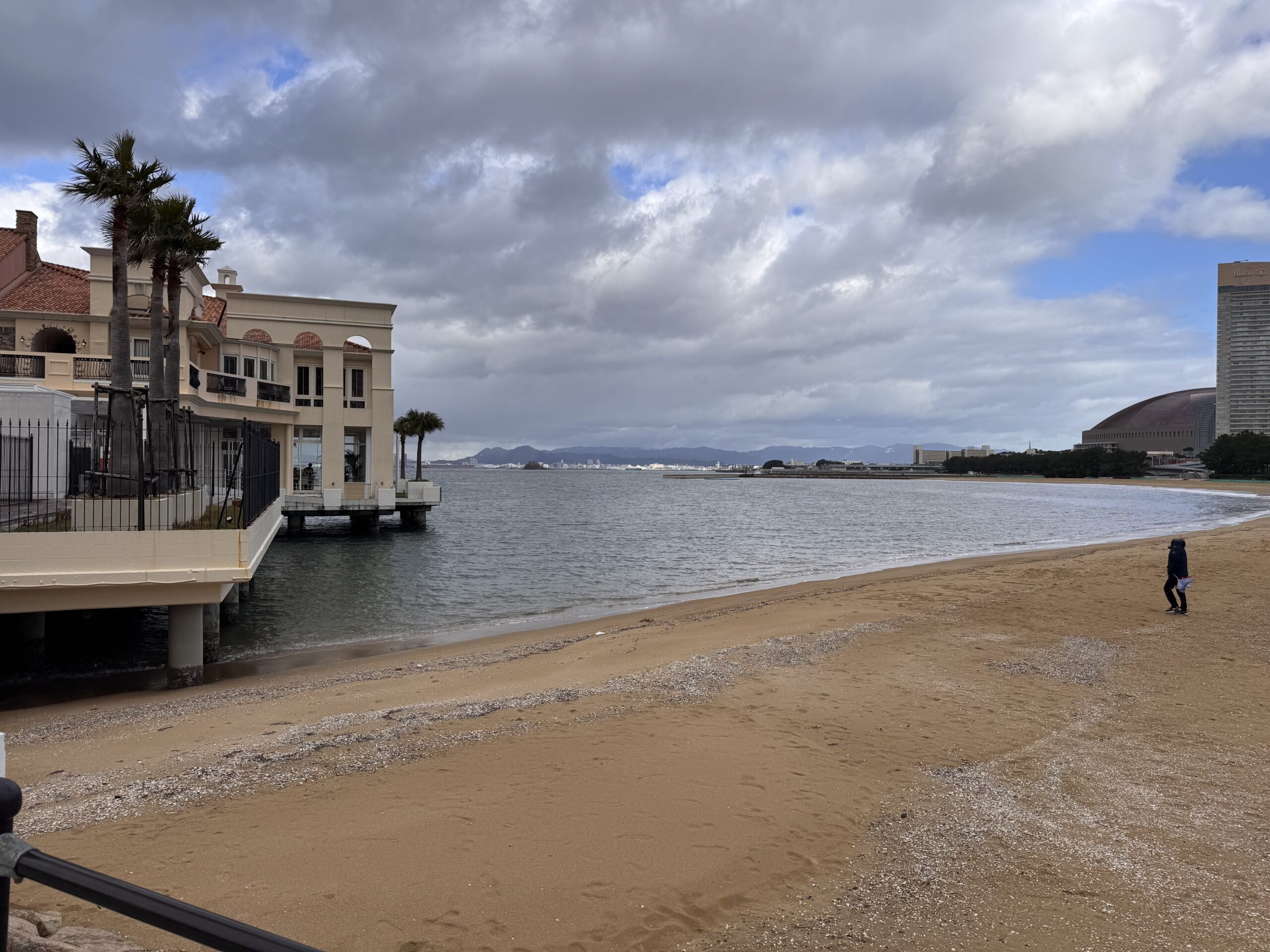 A view of Momochi Beach in Fukuoka, Japan, with sandy shores, seaside buildings, and Fukuoka Dome in the distance under a partly cloudy sky. (후쿠오카 모모치 해변 풍경 – 모래사장, 바닷가 건물, 멀리 보이는 후쿠오카 돔과 구름 낀 하늘)