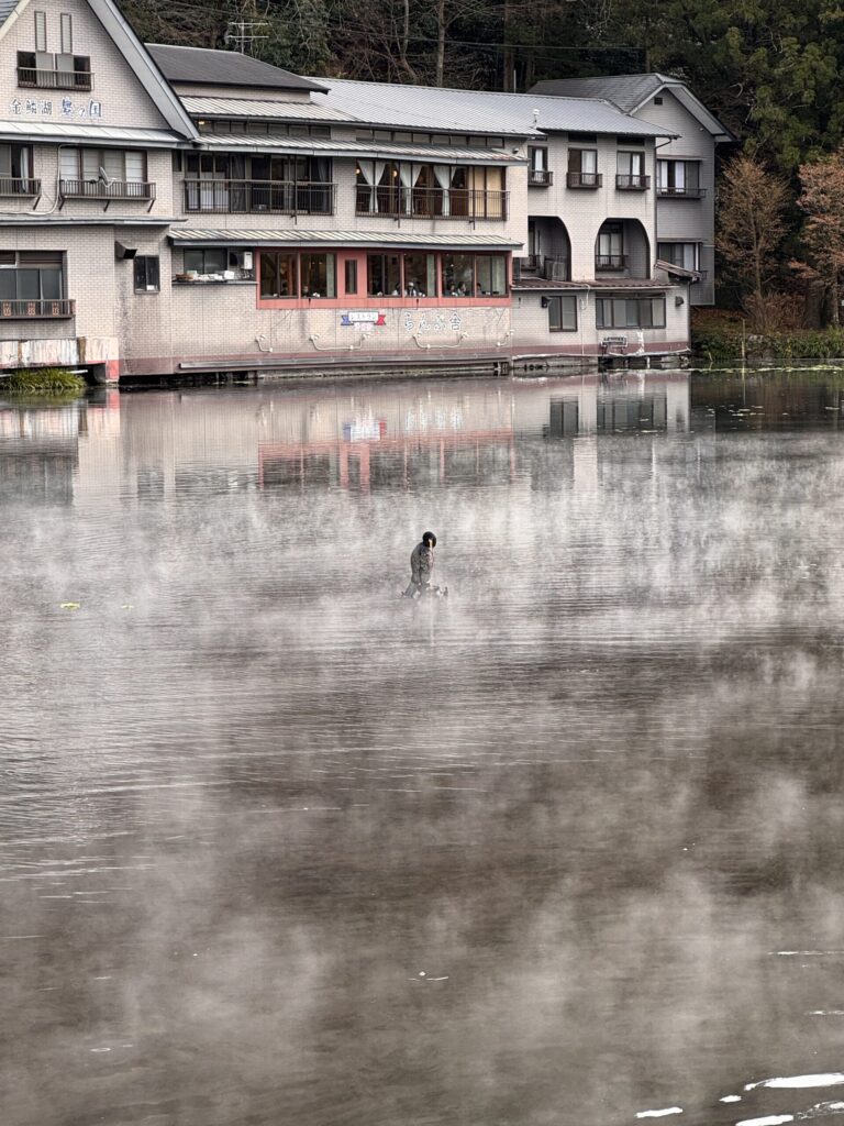 A person walking in a misty lake with steam rising from the water, with a building visible in the background.