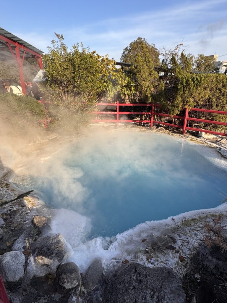 A steaming hot spring with bright blue water surrounded by rocks and red railings in a serene natural setting.