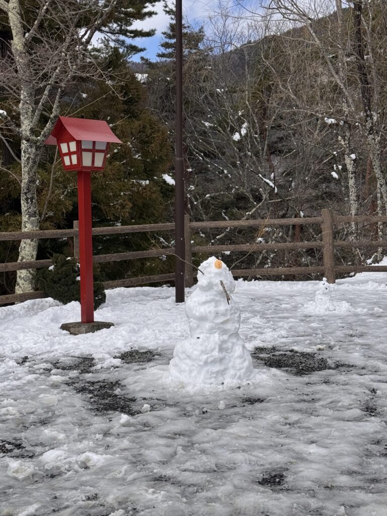  A snowman standing next to a red lantern in a snowy area, with a second snowman visible in the background.