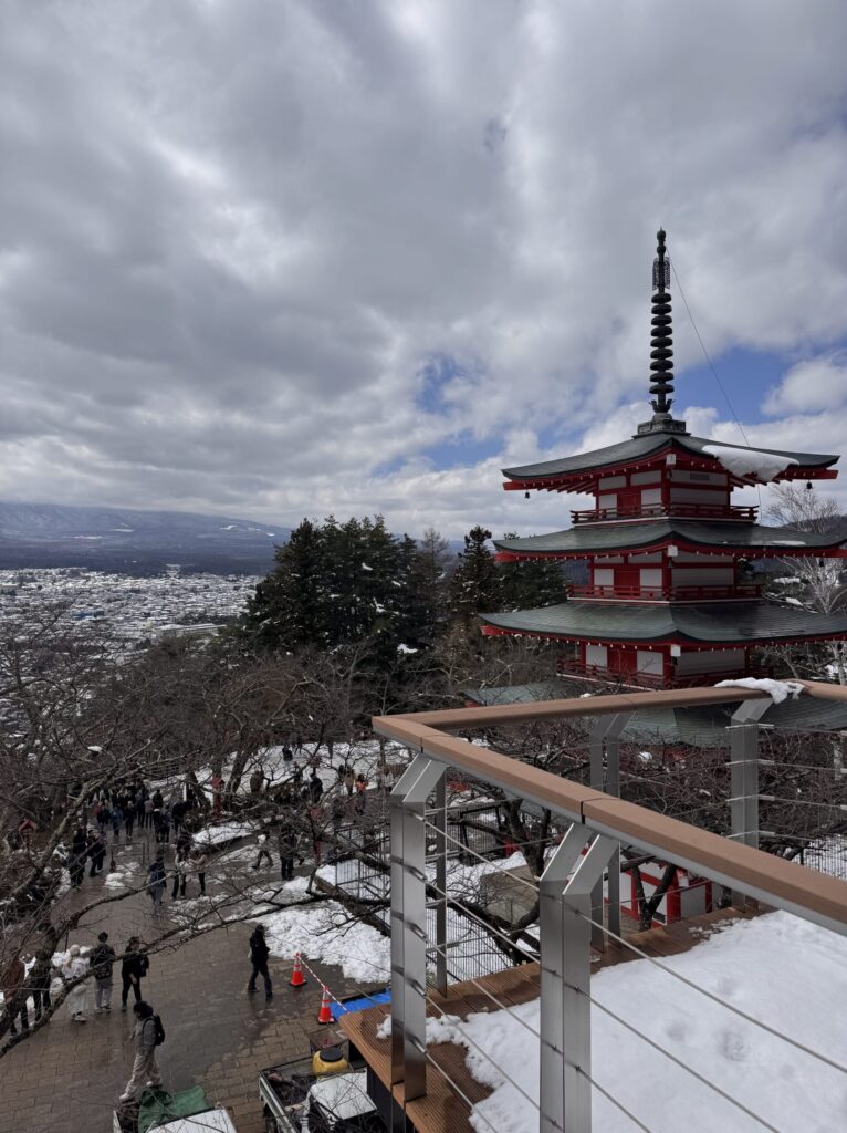A traditional five-story pagoda surrounded by snow-covered trees, with a cityscape in the background and a cloudy sky above.
