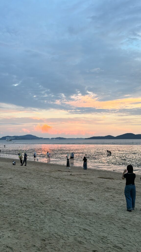 People walking and enjoying the view of the sea during sunset, with some engaging in activities on the wet sand as the sky glows in pink and orange hues.