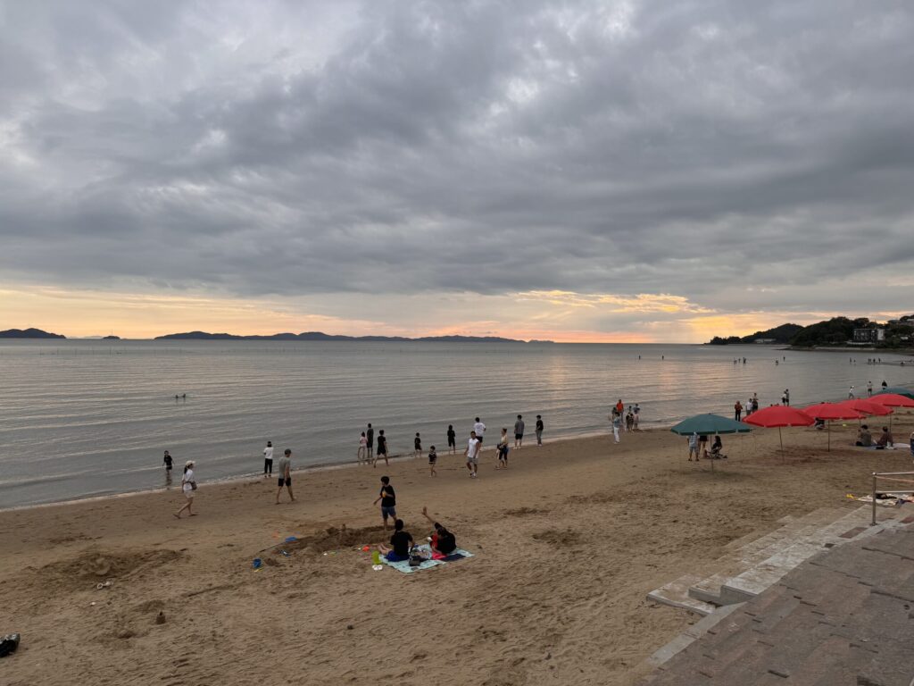 A beach scene at dusk, with people walking along the shore and a few umbrellas set up, with the sky showing a soft, orange glow.