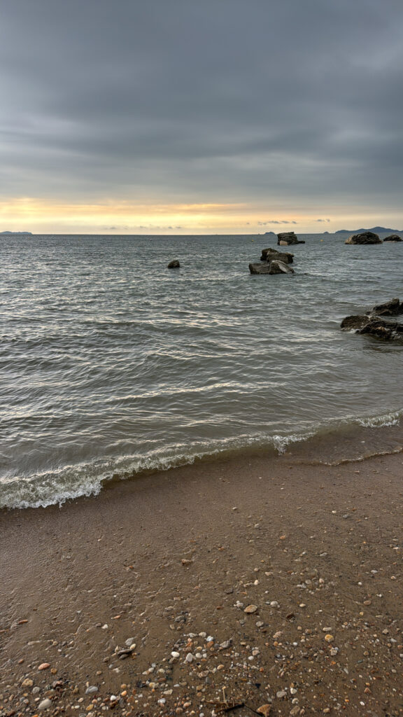 Close-up of the shoreline with gentle waves washing onto the sand, with rocky formations and mountains visible in the distance.