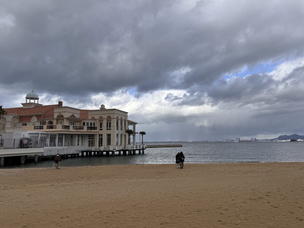 A tranquil beach scene with a view of a building on the left and two figures walking along the shoreline, with a cloudy sky and a hint of blue peeking through.