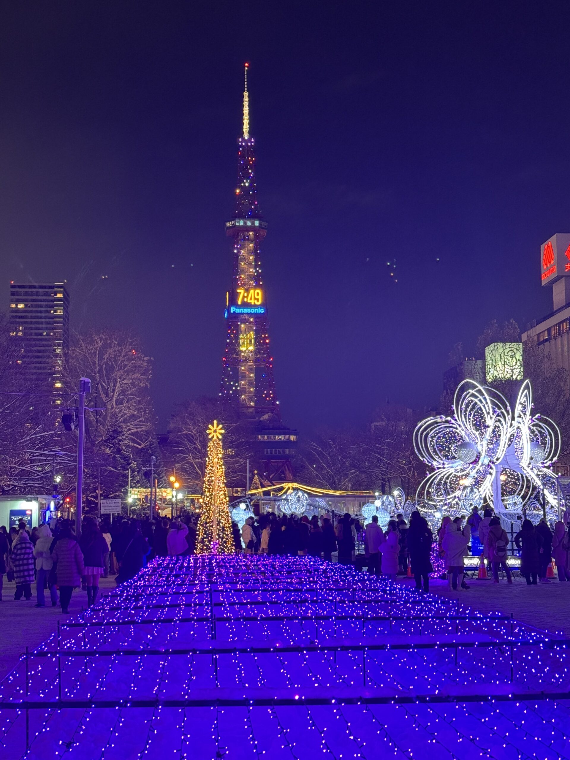 Sapporo TV Tower illuminated at night during the winter light festival, with glowing trees, Christmas decorations, and crowds enjoying the snowy evening. (겨울 빛 축제로 환하게 빛나는 삿포로 TV 타워와 크리스마스 장식, 눈 내린 저녁을 즐기는 사람들)