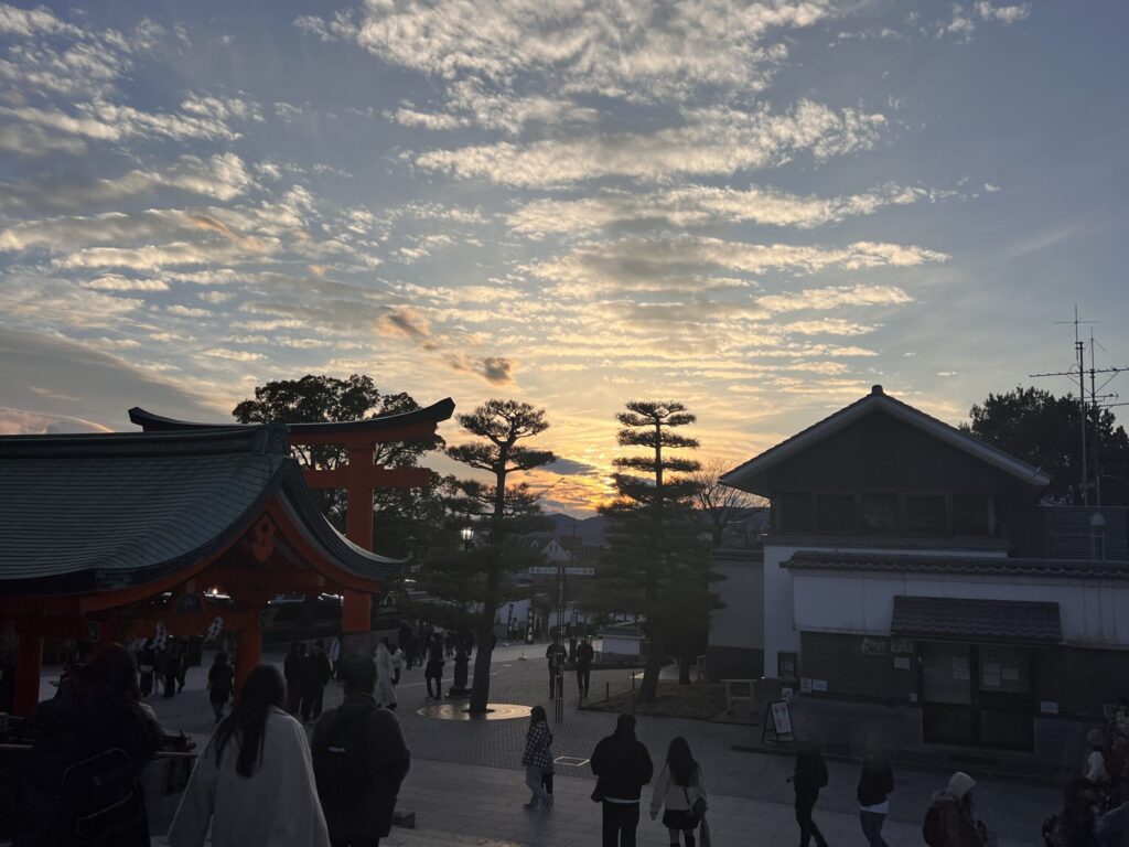 A group of people walking through the Fushimi Inari shrine area at sunset, with the iconic torii gates in the background.