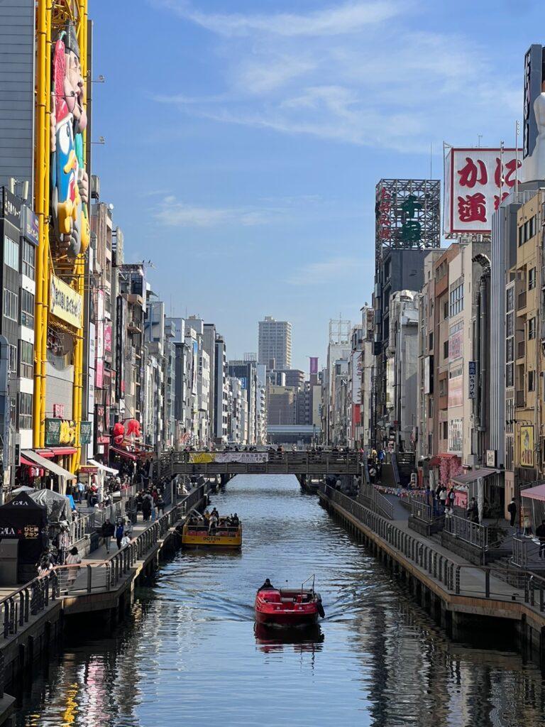 A view of the busy canal and buildings in Osaka, Japan, with a boat passing underneath a bridge and advertisements on the buildings.
