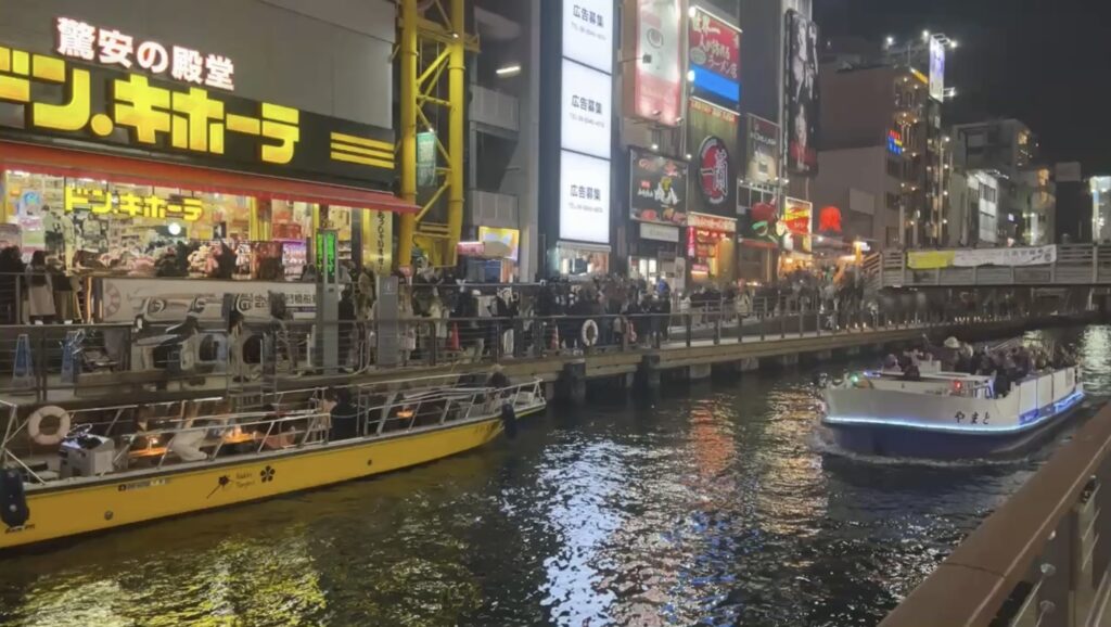 A view of the canal at night with boats docked and people walking along the waterfront in Osaka, illuminated by bright lights and neon signs.