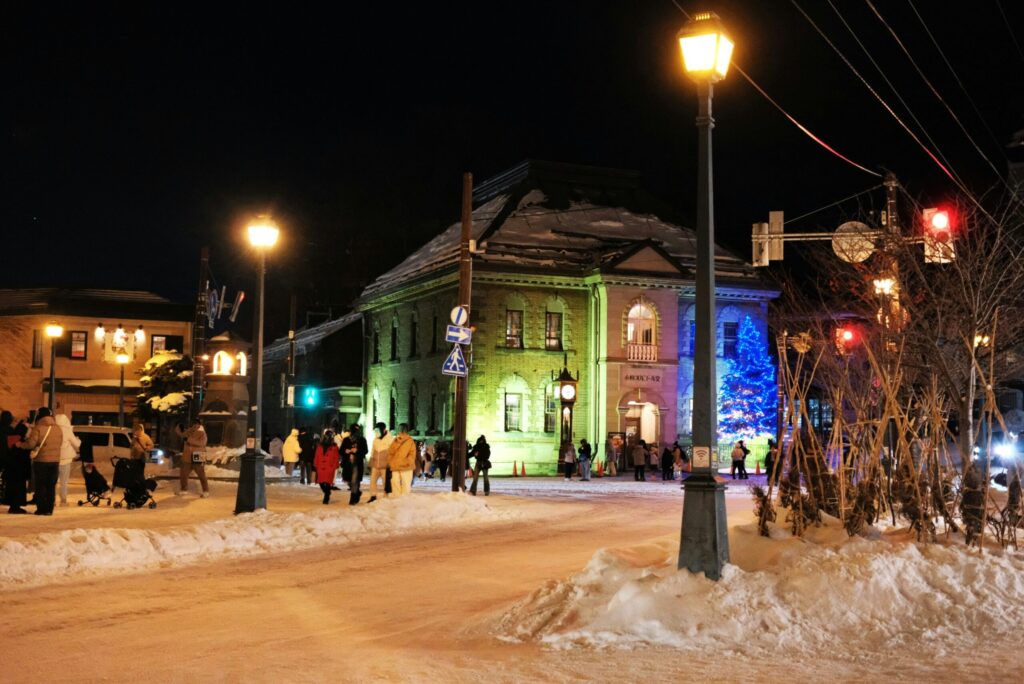 A winter night scene with a green-lit building, snow-covered streets, and people walking along the snowy road illuminated by streetlights.