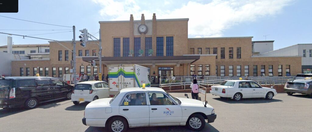 A daytime view of a brick train station building with a clock, with cars and people moving around outside.