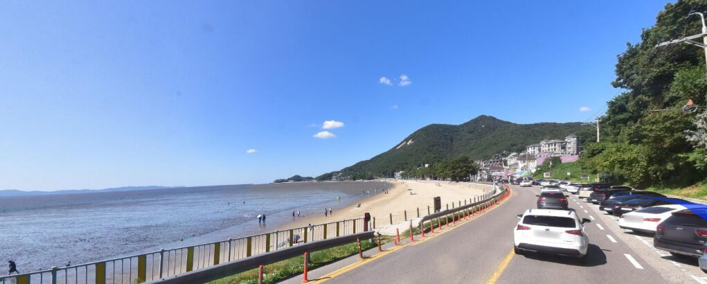 A clear blue sky over a beach with a mountain in the background, with people walking along the beach and cars on a nearby road.