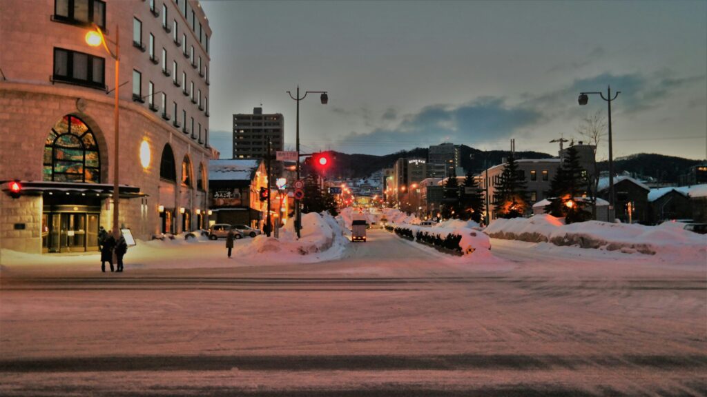 A snowy street scene with a view of buildings, snow piles, and streetlights illuminating the path ahead, creating a cozy winter atmosphere.