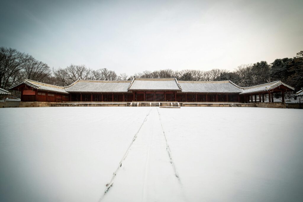 A traditional Korean building covered in snow, with a wide open space in front of it and bare trees around.