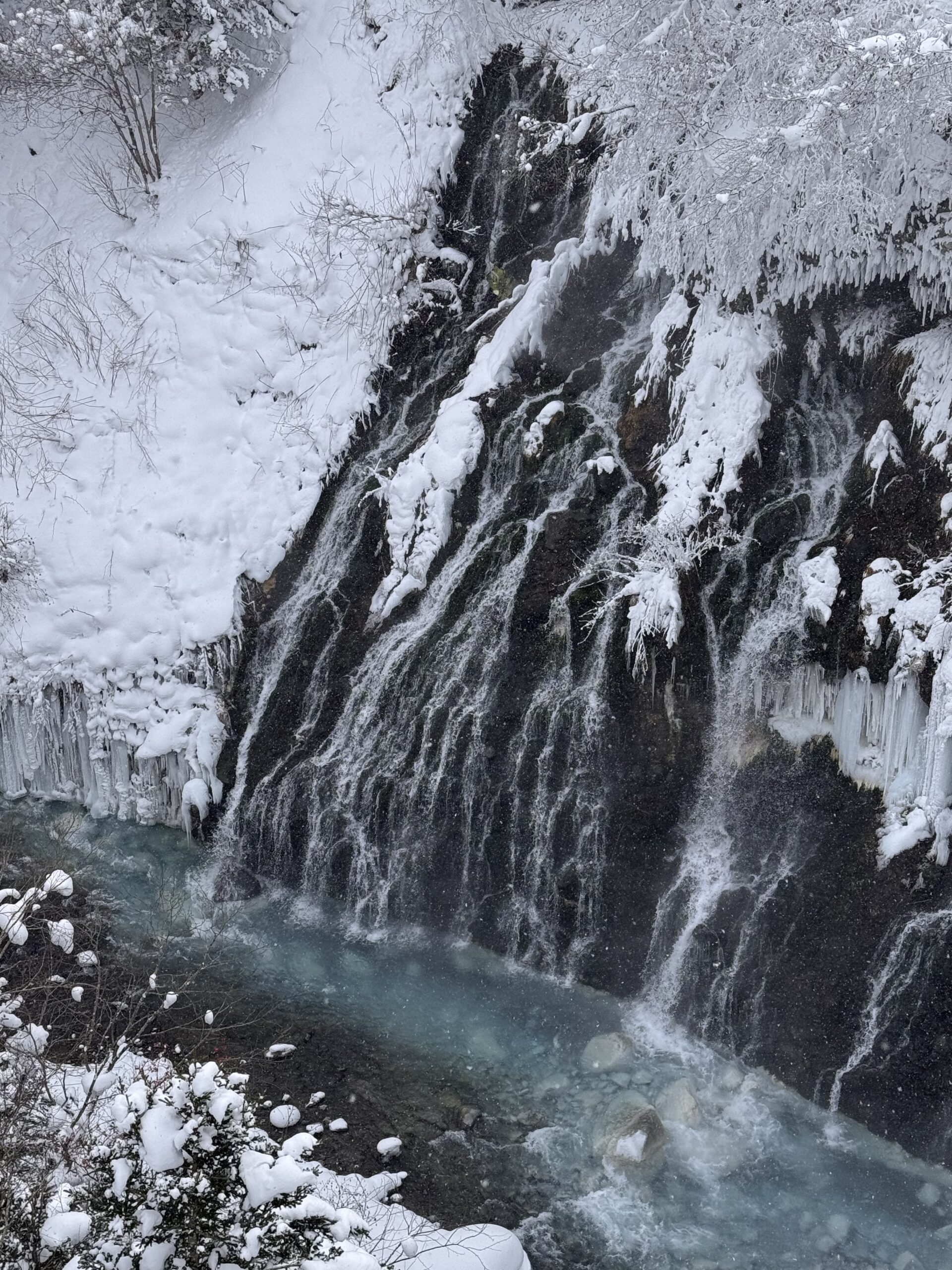 EN: A frozen waterfall flowing through a snowy forest in winter. KR: 겨울 숲 속, 눈과 얼음으로 뒤덮인 폭포의 모습.