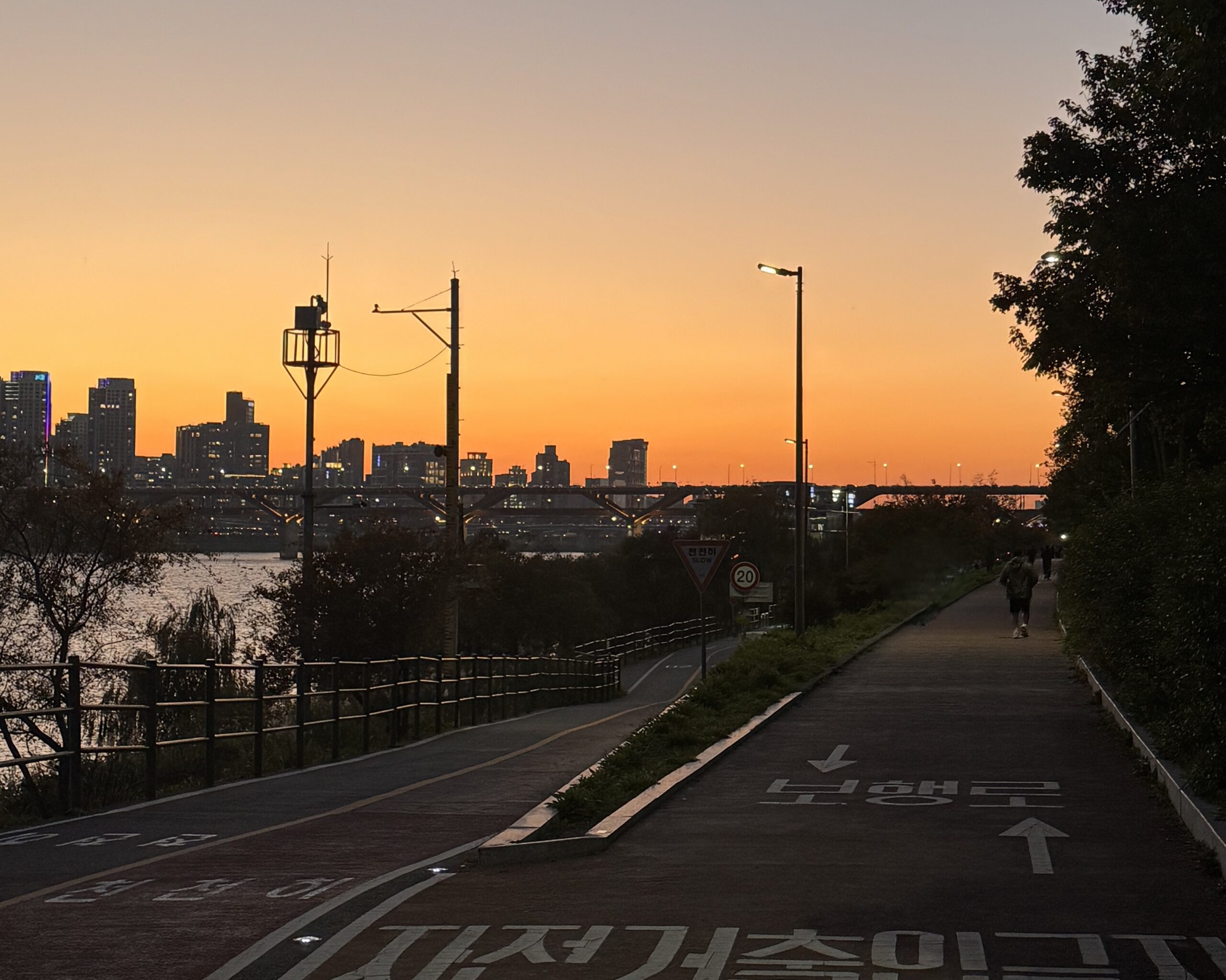 Sunset walkway by the Han River with Seoul skyline in the background. 한강 산책길 위로 석양과 서울 스카이라인이 펼쳐진 풍경.