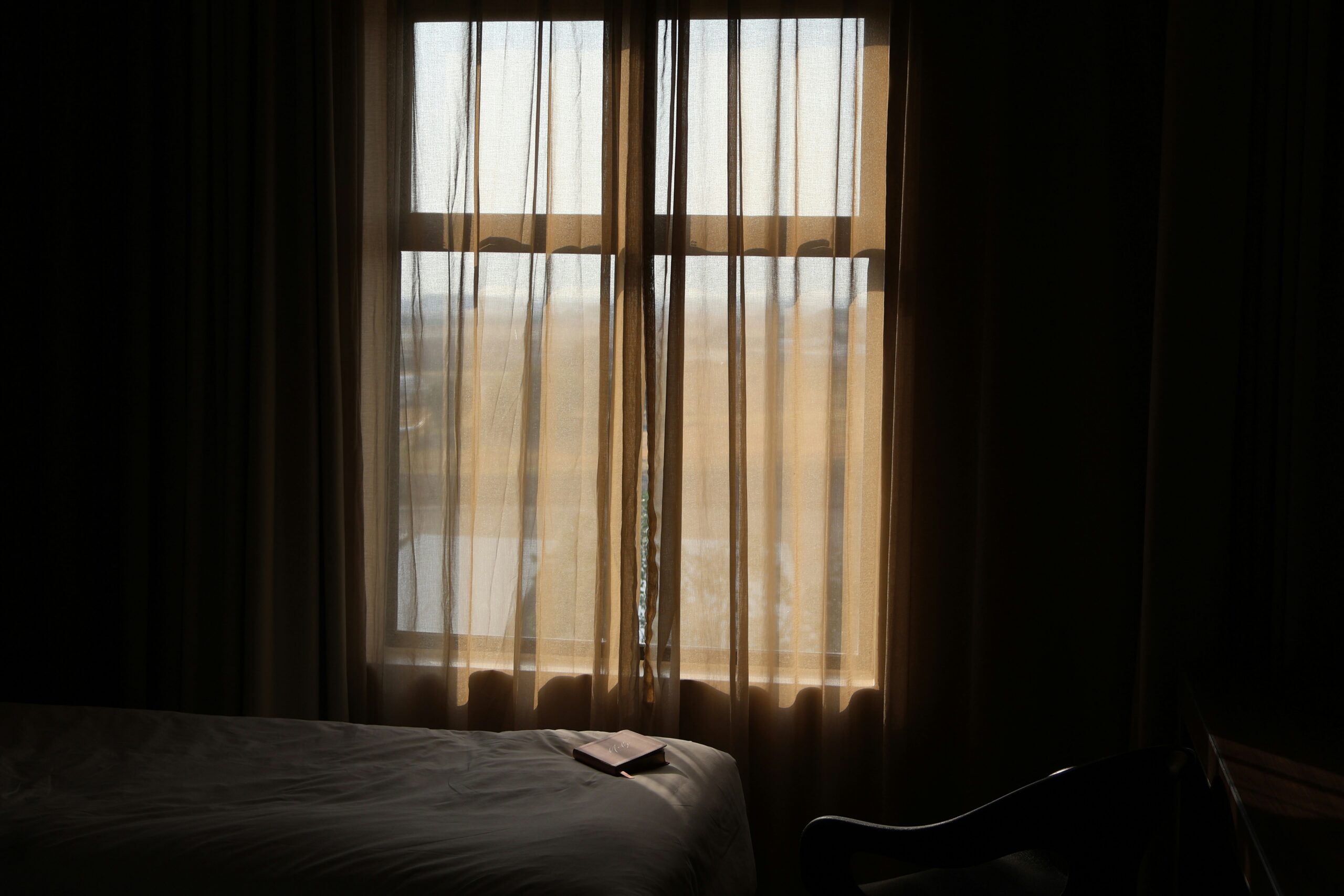KR: 커튼 사이로 빛이 들어오는 방, 침대 위에 놓인 노트 EN: A quiet room with soft light through the curtains and a notebook on the bed